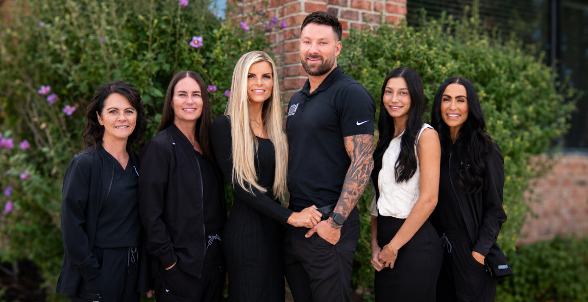 Six people, including a blonde woman and a bearded man in the center, standing together outdoors in front of green hedges and a brick wall, dressed mostly in black.